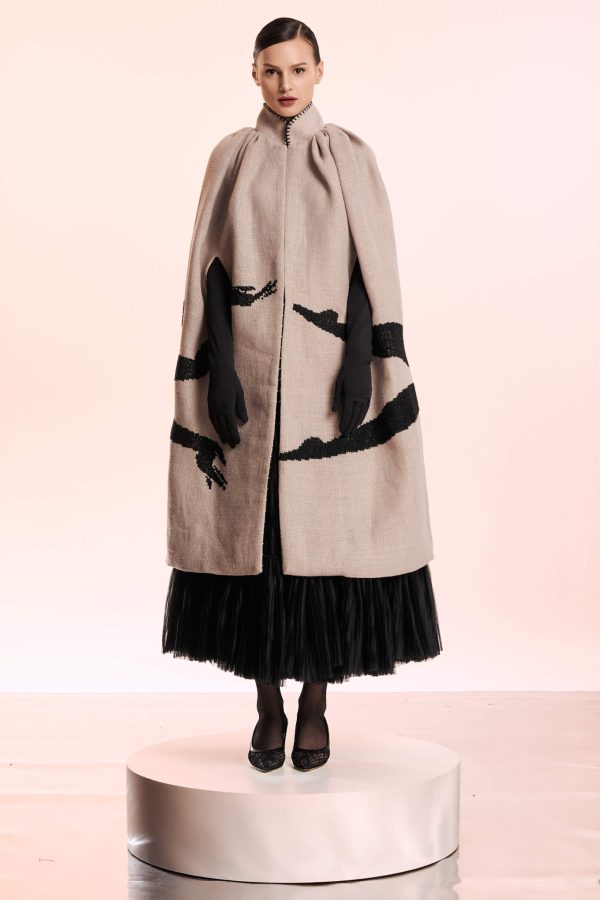 This garment cut from hep has a very distinctive gray-lila color. The silhouette that is hand embroidered looks as if it embraces the body wearing this cape. The collar has a black detail also sewn by hand.