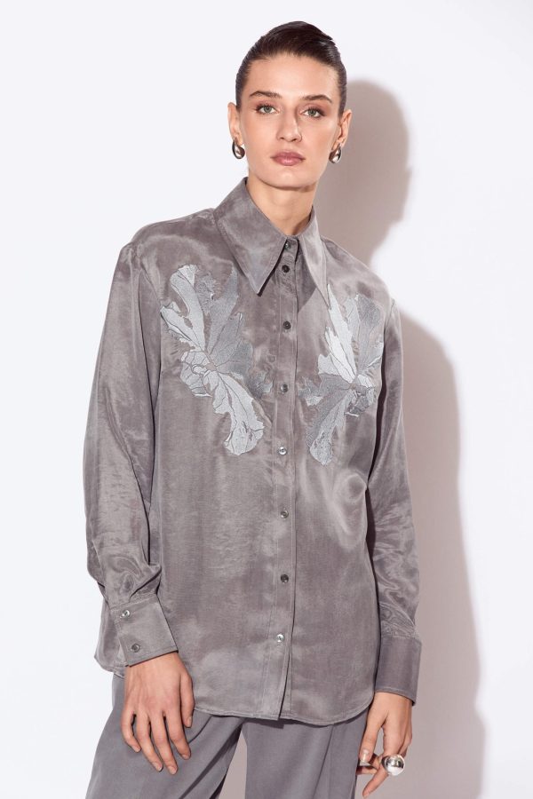 A free volume shirt with a straight silhouette, a curly bottom line, and a length below the hip line. The shoulder line is lowered and it has a symmetrical embroidery on the chest area.