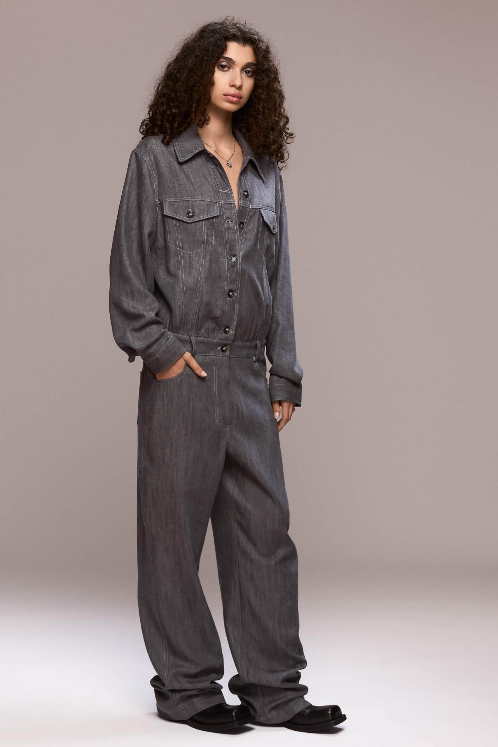 This gray demin boilersuit is a long-sleeved, full-length garment with a straight leg cut, has a front closure with buttons and zipper for the trousers. It features patch pockets on both sides of the chest and 4 functional ones, two in front and other two on the back of the trousers. Its fit is comfortable yet flattering, making it a versatile wardrobe piece. Wear it on it's own or with a contrasting belt.