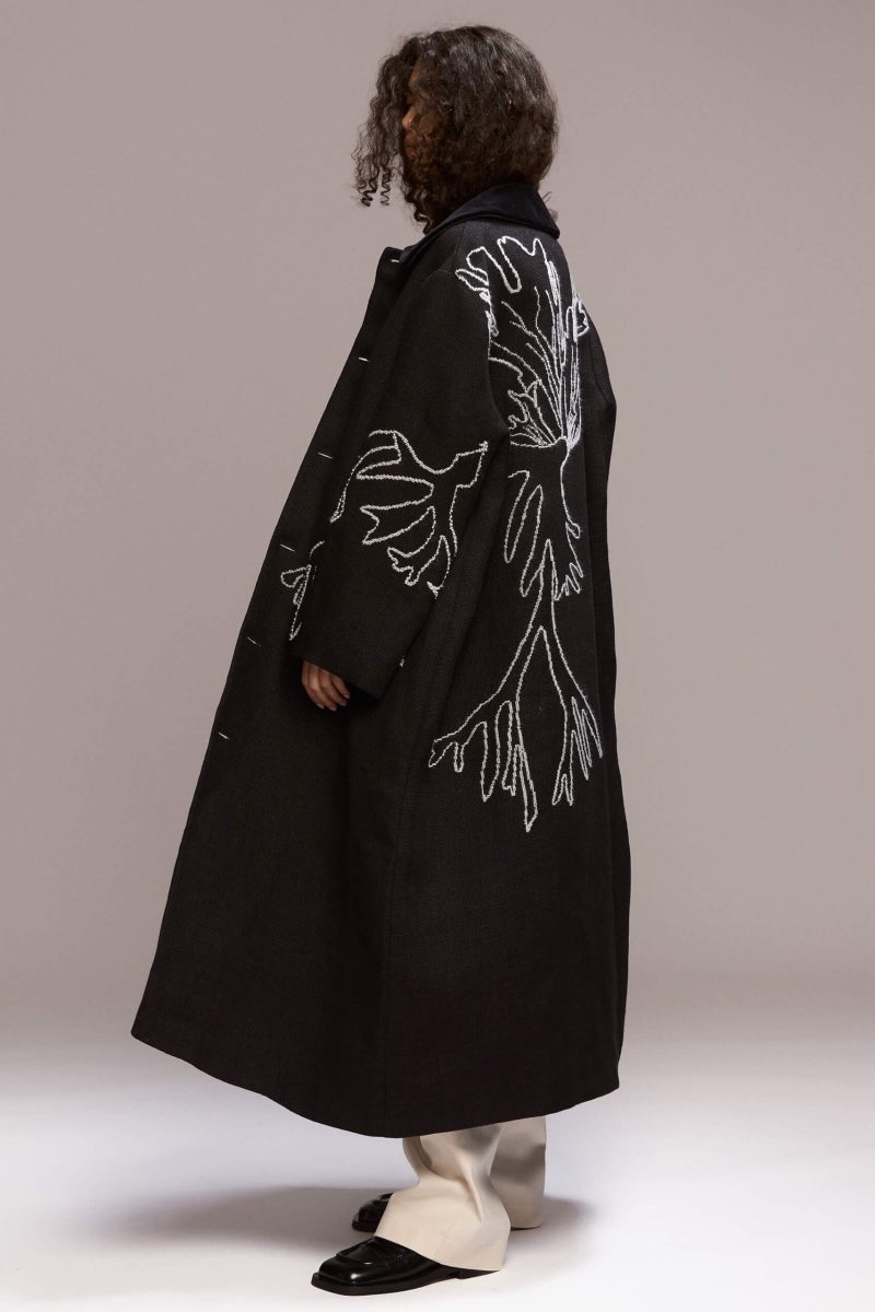 This long black coat features a relaxed, oversized fit with front button closure and a velvet collar. Crafted from hemp, it extends to a maxi lenght. A distinctive white manual embroidery adorns the back and left front side of the coat as well as the hand, adding a unique design element.