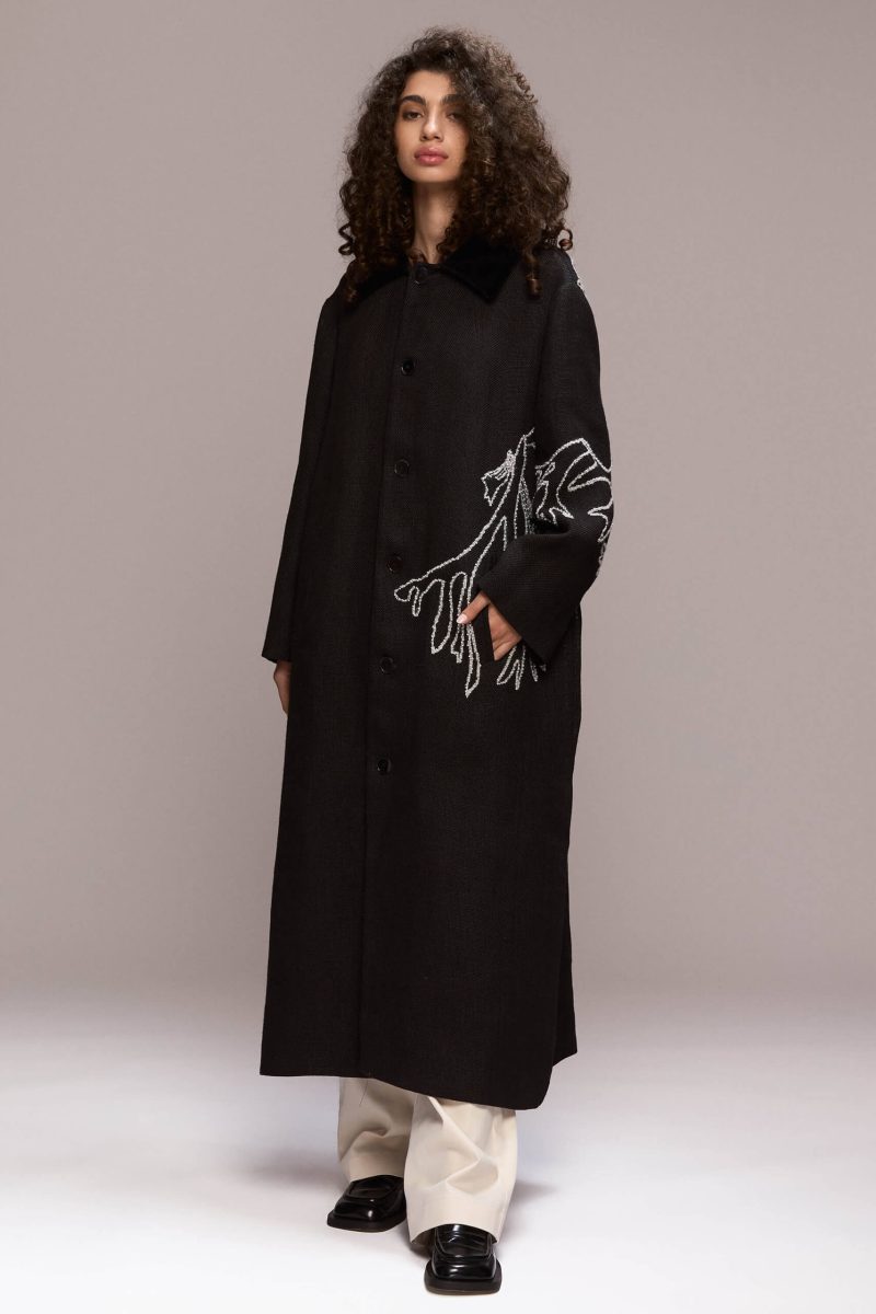 This long black coat features a relaxed, oversized fit with front button closure and a velvet collar. Crafted from hemp, it extends to a maxi lenght. A distinctive white manual embroidery adorns the back and left front side of the coat as well as the hand, adding a unique design element.