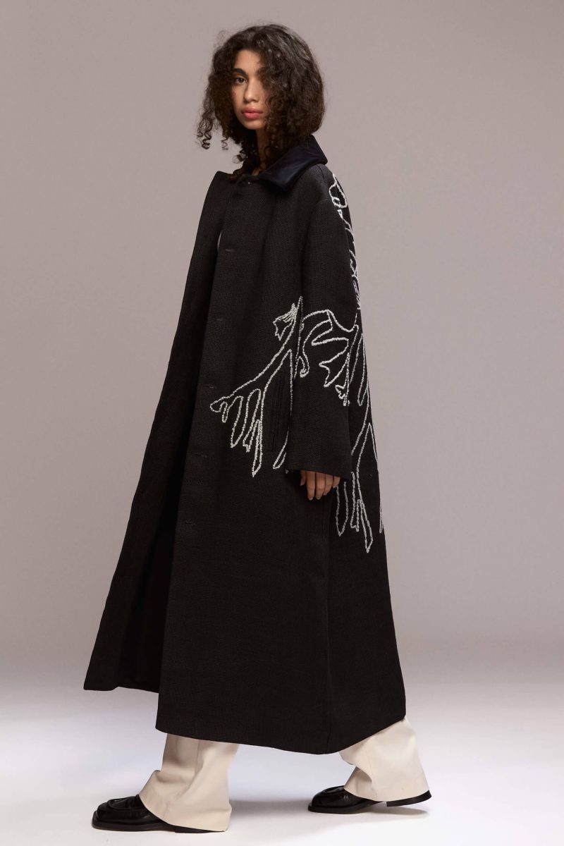 This long black coat features a relaxed, oversized fit with front button closure and a velvet collar. Crafted from hemp, it extends to a maxi lenght. A distinctive white manual embroidery adorns the back and left front side of the coat as well as the hand, adding a unique design element.