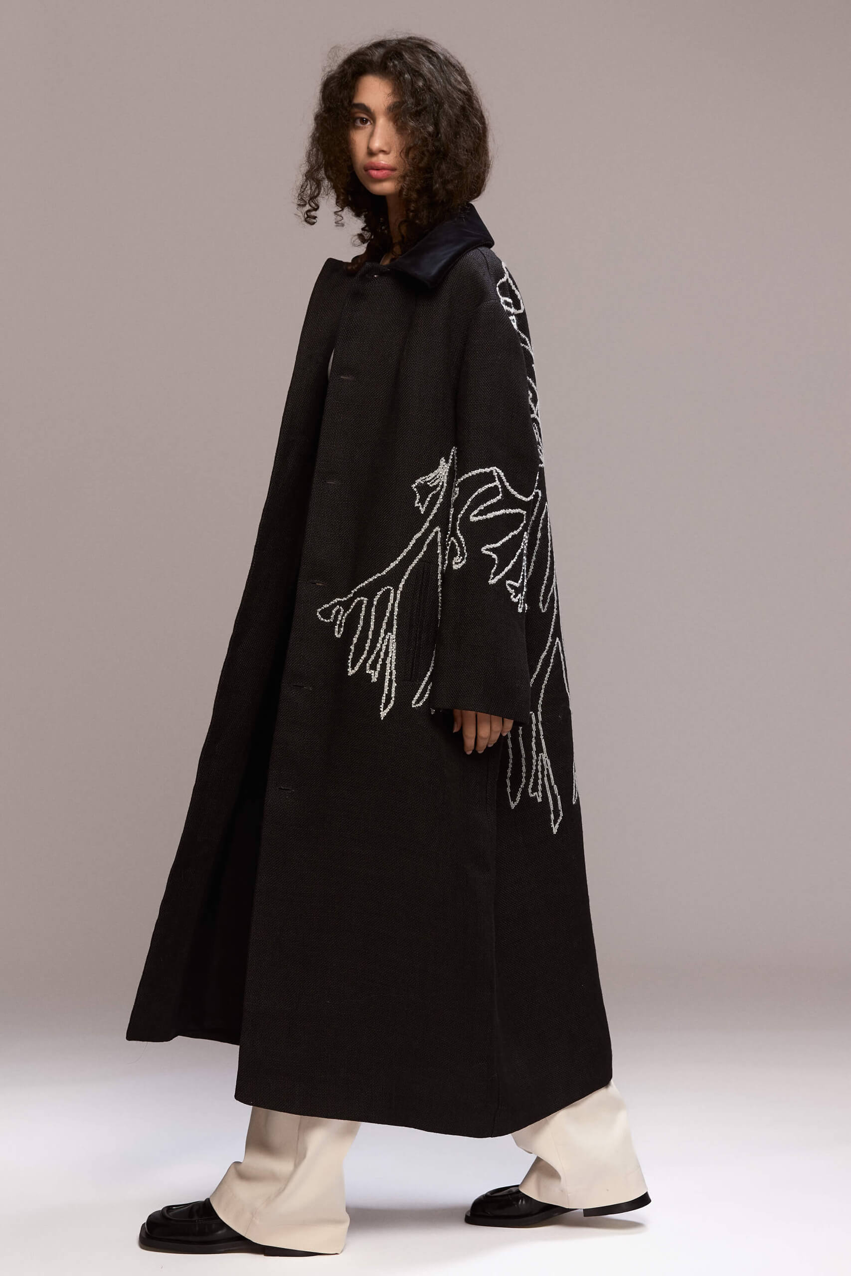 This long black coat features a relaxed, oversized fit with front button closure and a velvet collar. Crafted from hemp, it extends to a maxi lenght. A distinctive white manual embroidery adorns the back and left front side of the coat as well as the hand, adding a unique design element.