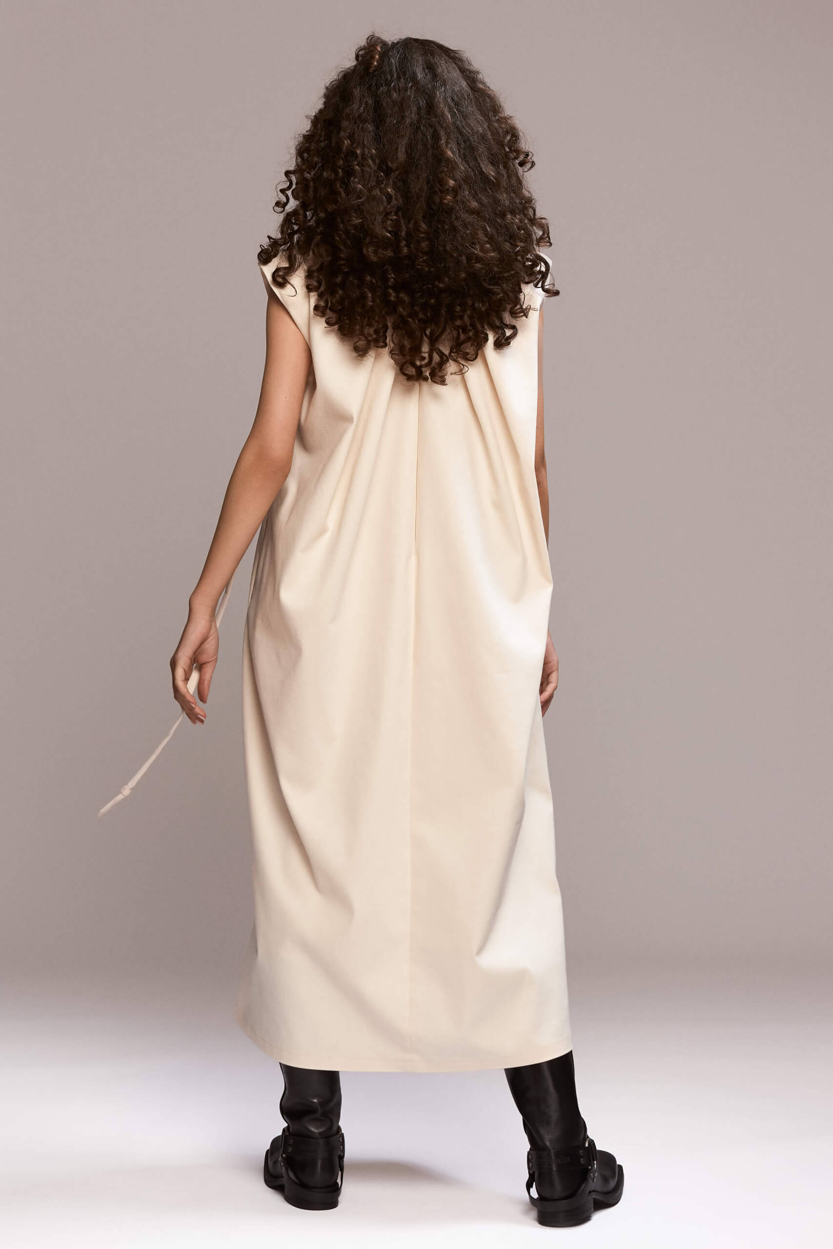 Off-white sleeveless, loose-fit maxi dress designed to flow on the body, with beautiful back pleats and structured padded shoulders. The dress includes a removable self-tie belt to cinch the waist and side pockets.