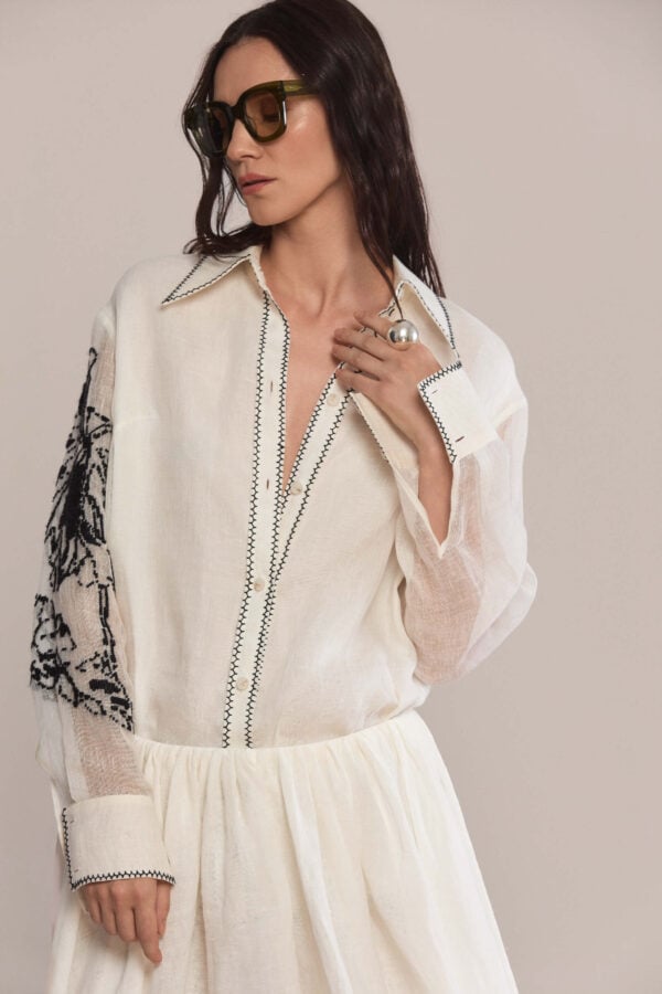 Crafted from 100% linen, this shirt combines timeless elegance with subtle detailing. It features a classic collar and sleek black topstitching along the placket, collar, and hem, while bold black embroidery on the left sleeve adds an artistic, standout touch.