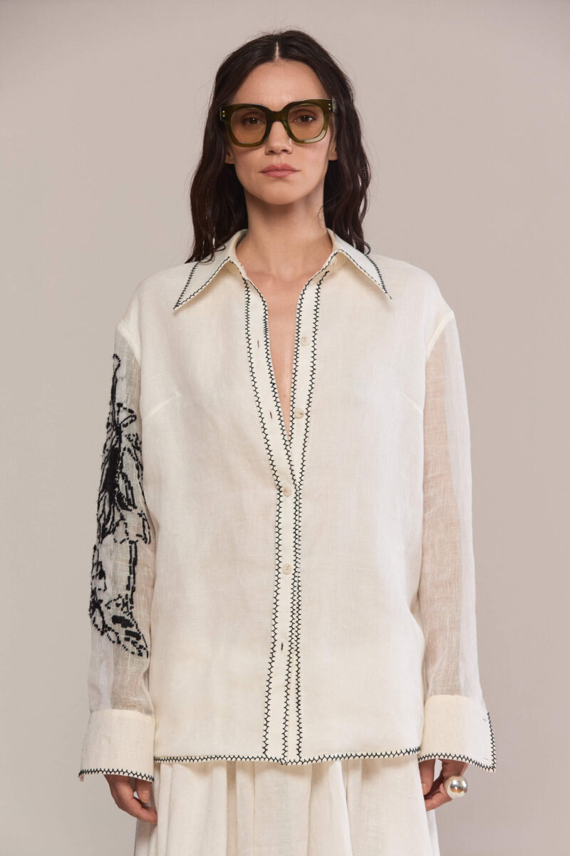 Crafted from 100% linen, this shirt combines timeless elegance with subtle detailing. It features a classic collar and sleek black topstitching along the placket, collar, and hem, while bold black embroidery on the left sleeve adds an artistic, standout touch.