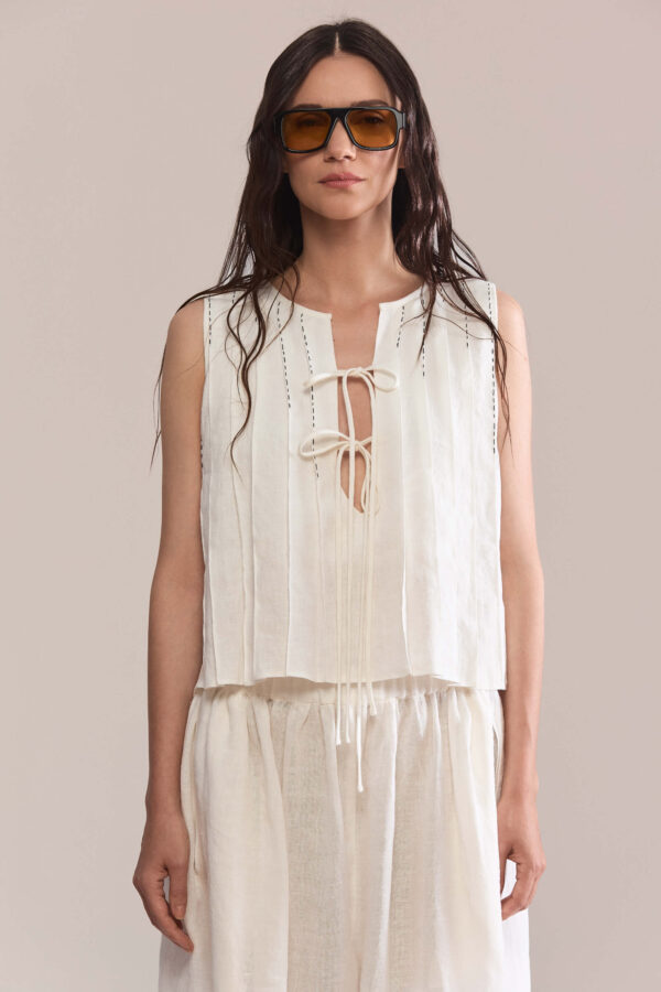 A relaxed sleeveless top with a soft V-neckline and delicate self-tie details. Vertical stitching and a slightly cropped hem add subtle character. Style it with the matching draped balloon pants or denim for an effortless warm-weather look.