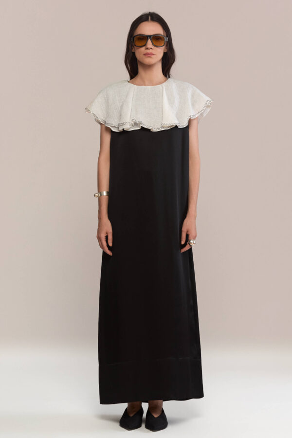 A floor-length black silk dress with a sleek, straight silhouette. The dramatic oversized white ruffled linen collar falls elegantly over the shoulders, offering a bold contrast and refined monochrome appeal. This dress is a true statement.