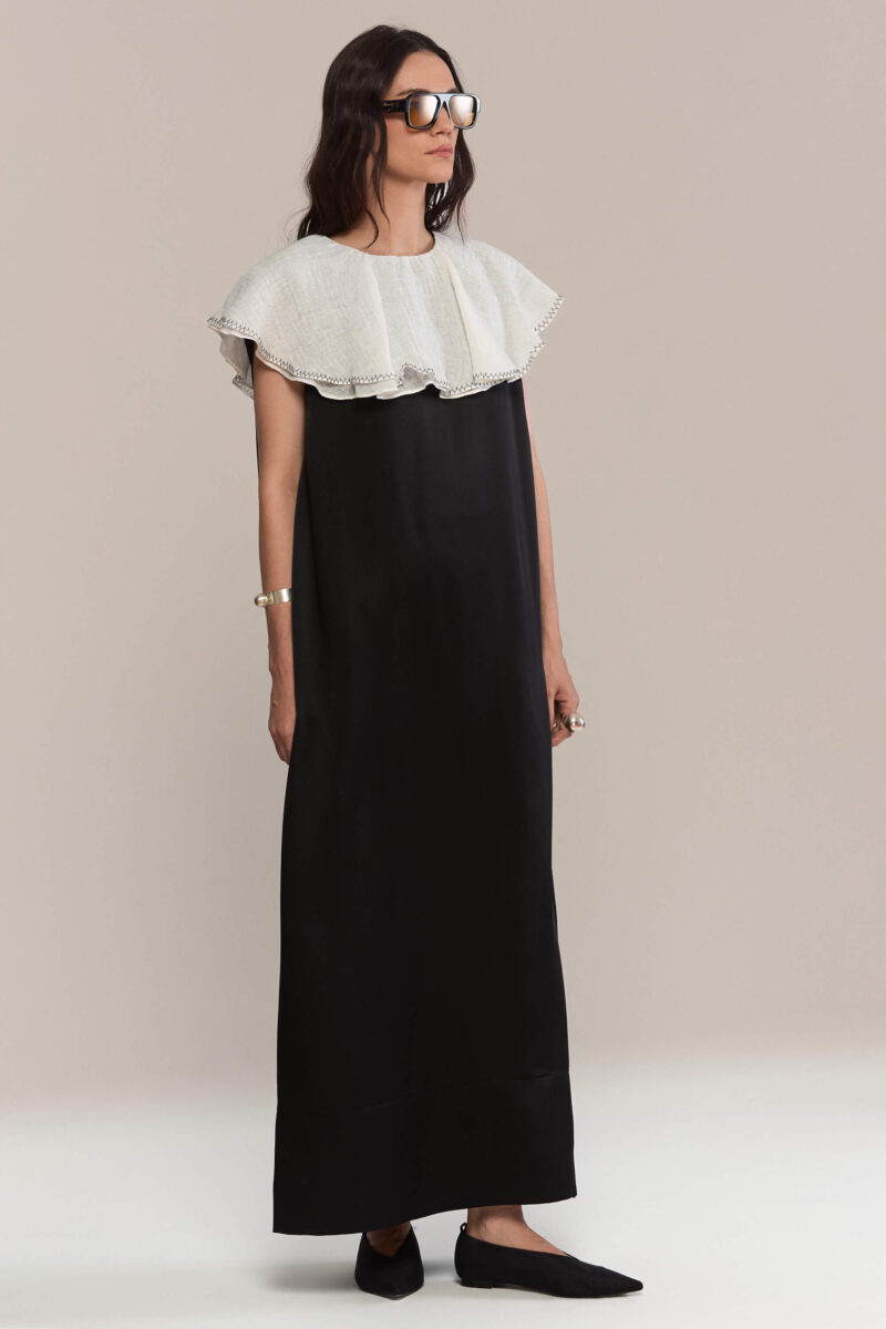 A floor-length black silk dress with a sleek, straight silhouette. The dramatic oversized white ruffled linen collar falls elegantly over the shoulders, offering a bold contrast and refined monochrome appeal. This dress is a true statement.