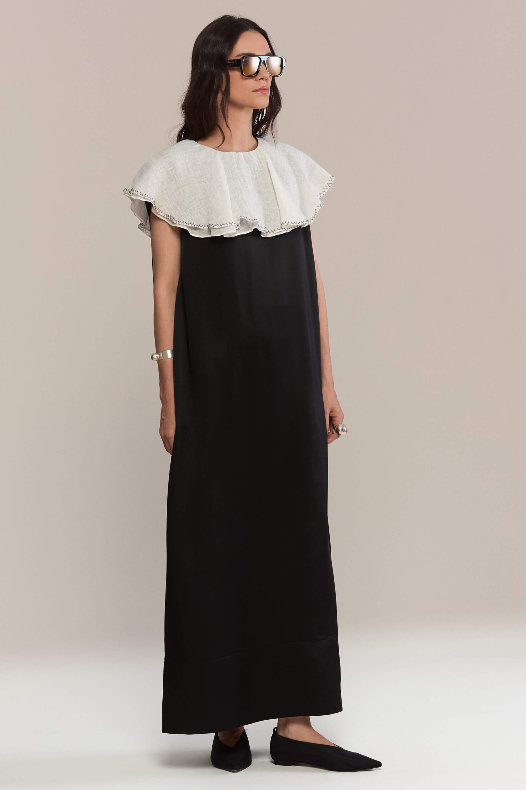 A floor-length black silk dress with a sleek, straight silhouette. The dramatic oversized white ruffled linen collar falls elegantly over the shoulders, offering a bold contrast and refined monochrome appeal. This dress is a true statement.